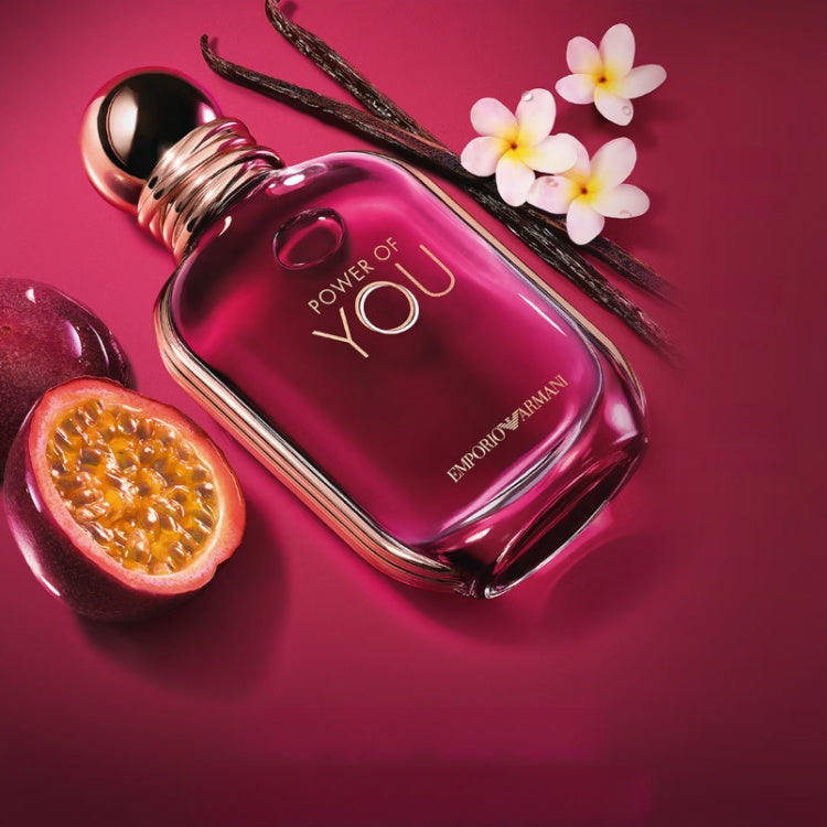 The power of you armani original perfume bottle displayed next to fresh passion fruit slices, vanilla beans, and white blossoms on a deep pink background.