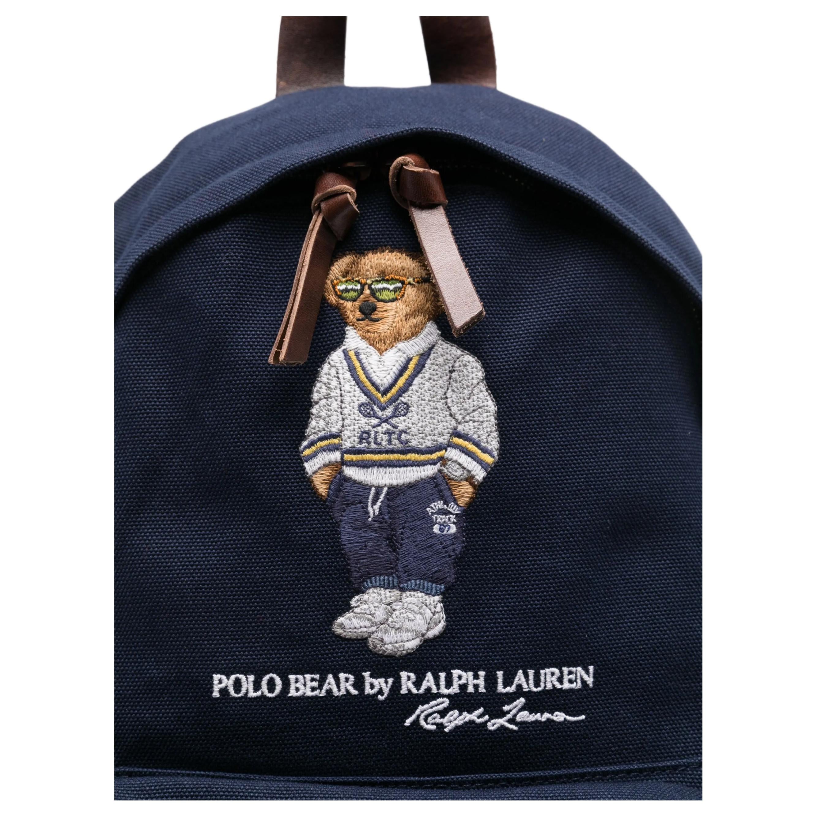 Close-up of the iconic embroidered Polo Bear wearing a sweater and glasses on an original navy Ralph Lauren backpack with brown leather zipper pulls.

لقطة قريبة لتطريز دب بولو الشهير الذي يرتدي سترة ونظارات على حقيبة ظهر رالف لورين أصلية باللون الكحلي مع أشرطة سحاب جلدية بنية.
