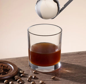Close up of metal ice tongs dropping a large, clear ice ball from an ice maker machine into a glass filled with a cold beverage.