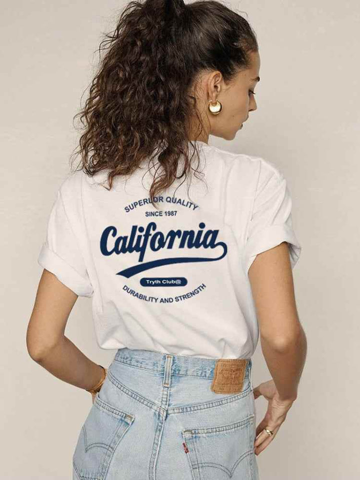 Woman wearing a white California Club casual cotton t-shirt with rolled sleeves and blue jeans, showing back print design.