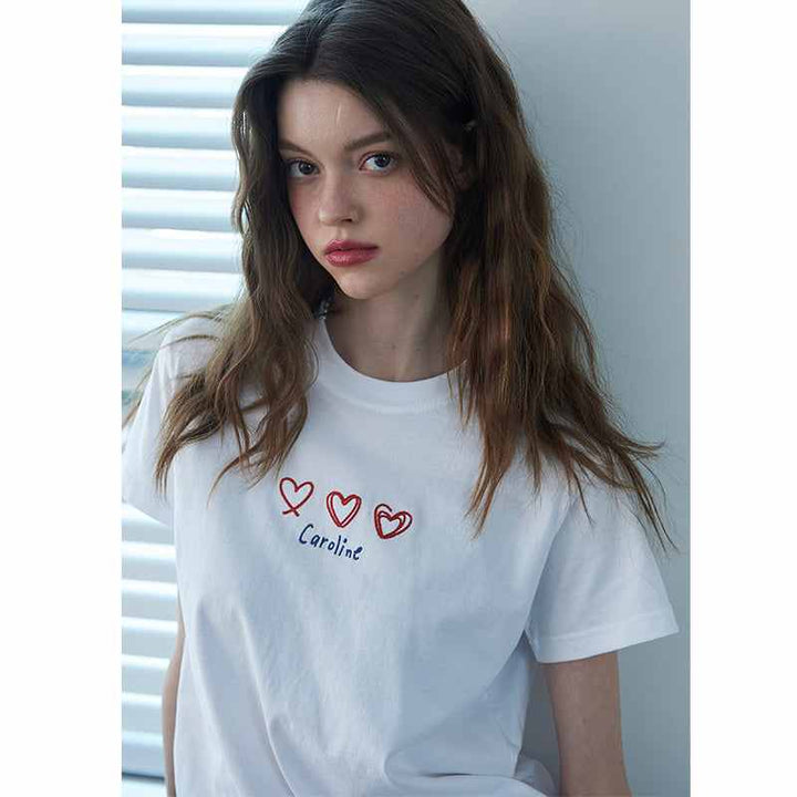 فتاة ترتدي تيشيرت Caroline Hearts أبيض ناعم مع تطريز ثلاث قلوب وكلمة Caroline بألوان حمراء</s>
