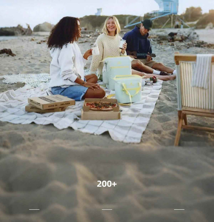 Three friends enjoying a beach picnic with Stanley insulated cups and coolers for hot and cold drinks