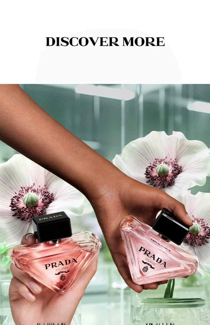 Two pink Prada Paradoxe perfume bottles held near white flowers with dark centers on reflective surface, showcasing luxury women's fragrance set.
