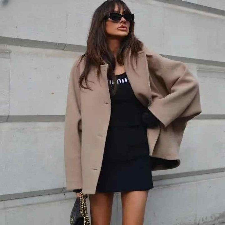 Woman wearing beige oversized winter coat over black dress and sunglasses outdoors against urban wall